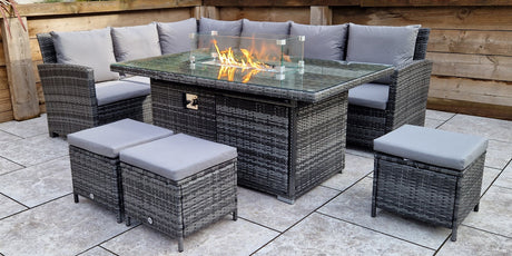 Modern grey garden dining set with cushioned seating and a central gas fire pit table, styled on a tiled patio with wooden fencing and decorative plants.